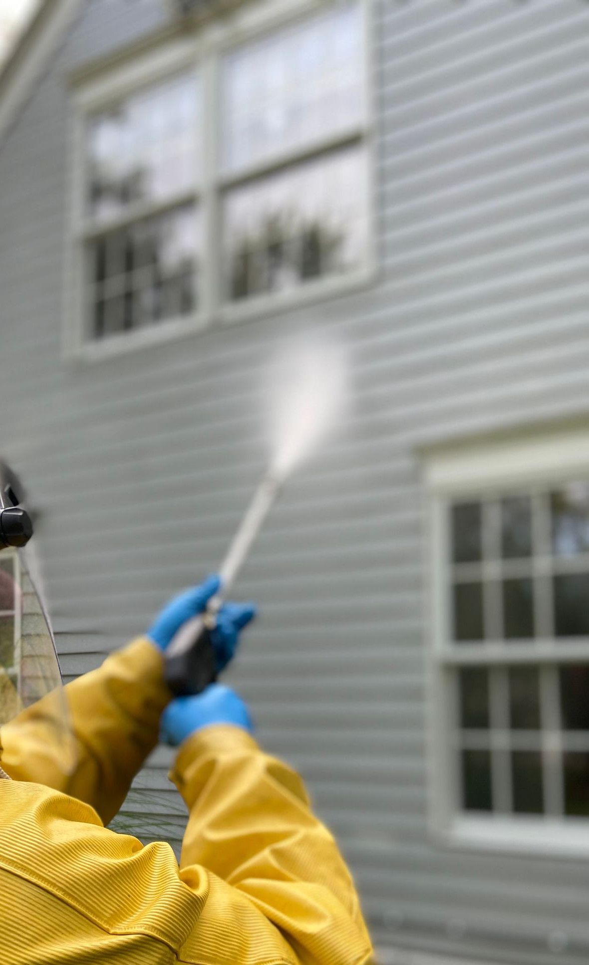 Person cleaning house siding with a pressure washer, wearing protective gear in a yellow raincoat.